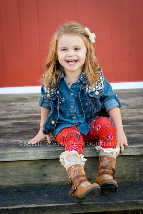 family photography session where little girl is sitting on wood deck in front of red barn smiling at the camera