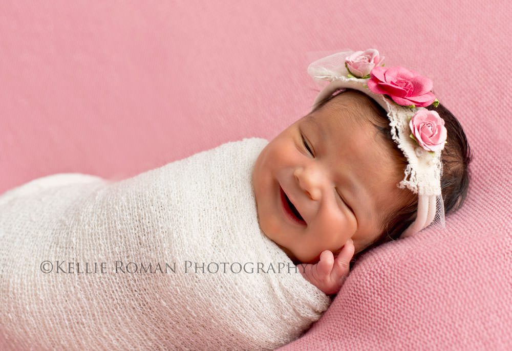 local photographers little infant girl in white swaddle blanket laying on back on pink blanket with  big smile wearing pink and white headband