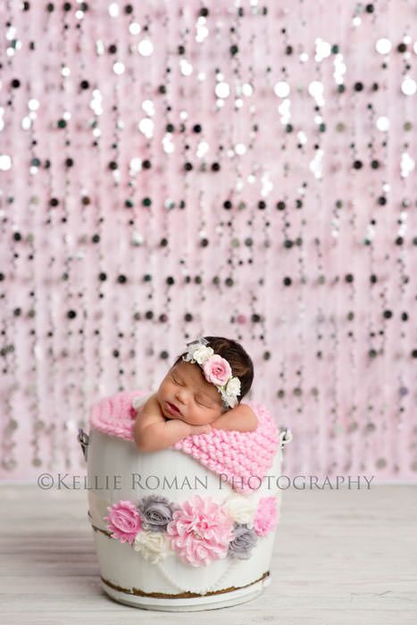 local photographers newborn girl posed in white bucket with floral sash  tied around it she's infant of a pink backdrop with silver beads hanging 