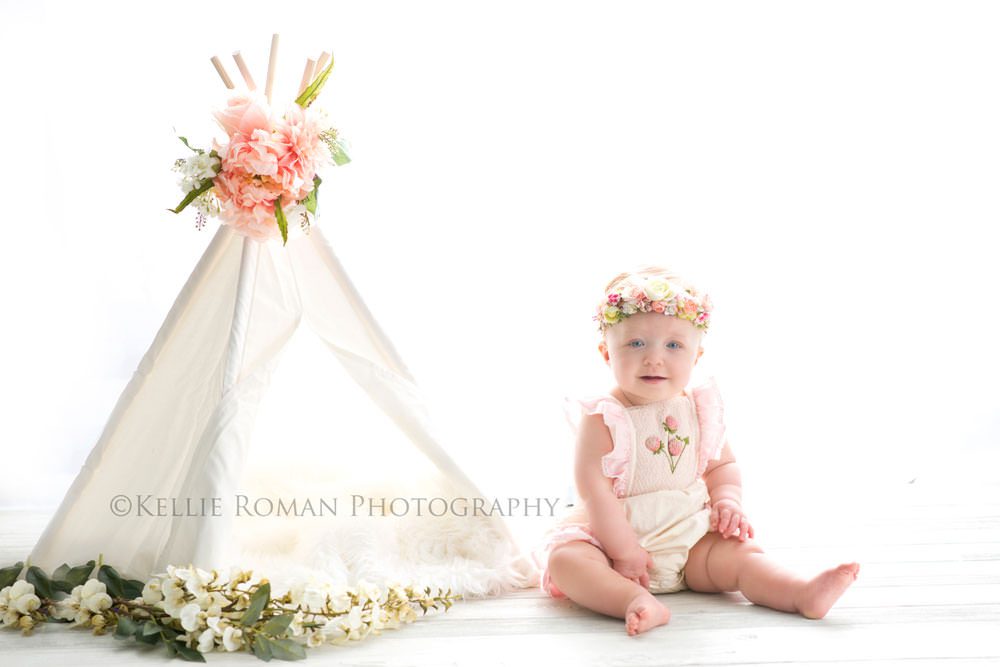 children photography one year old girl with pink romper on and flower crown sitting next to white teepee with flowers