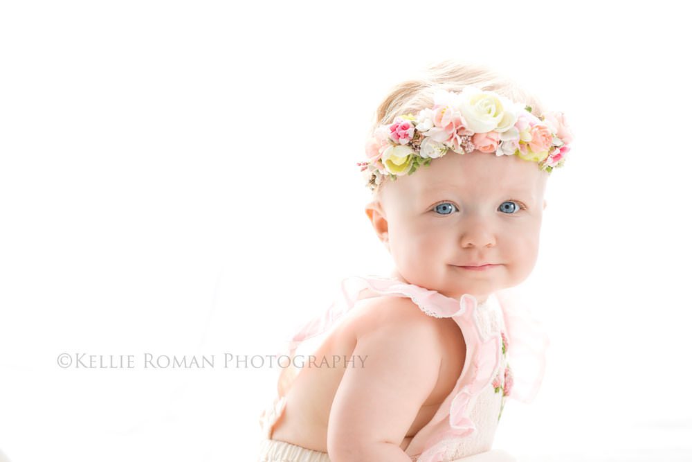 children photography one year old with big blue eyes and small smile looking at camera she has a pink romper and flower crown on