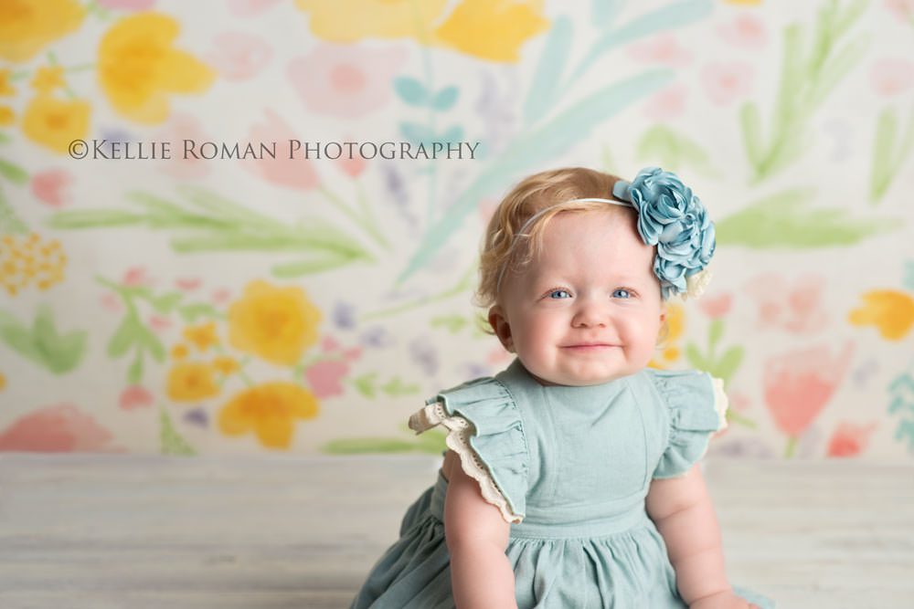 children photography one year old girl wearing a blue dress and blue headband sitting in front of floral backdrop with shades of yellow purple and pink 