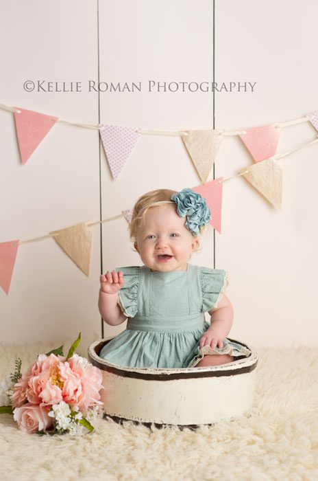 children photography one year old girl wearing a blue dress and blue headband sitting in ivory bucket with on top of an ivory rug with pink banner behind her she has pink flowers next to her