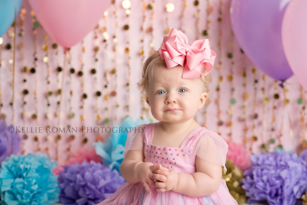 children photography one year old girt with birthday set up pink blue and purple balloons and pom poms and gold beads she's wearing a pink dress with big pink hair bow
