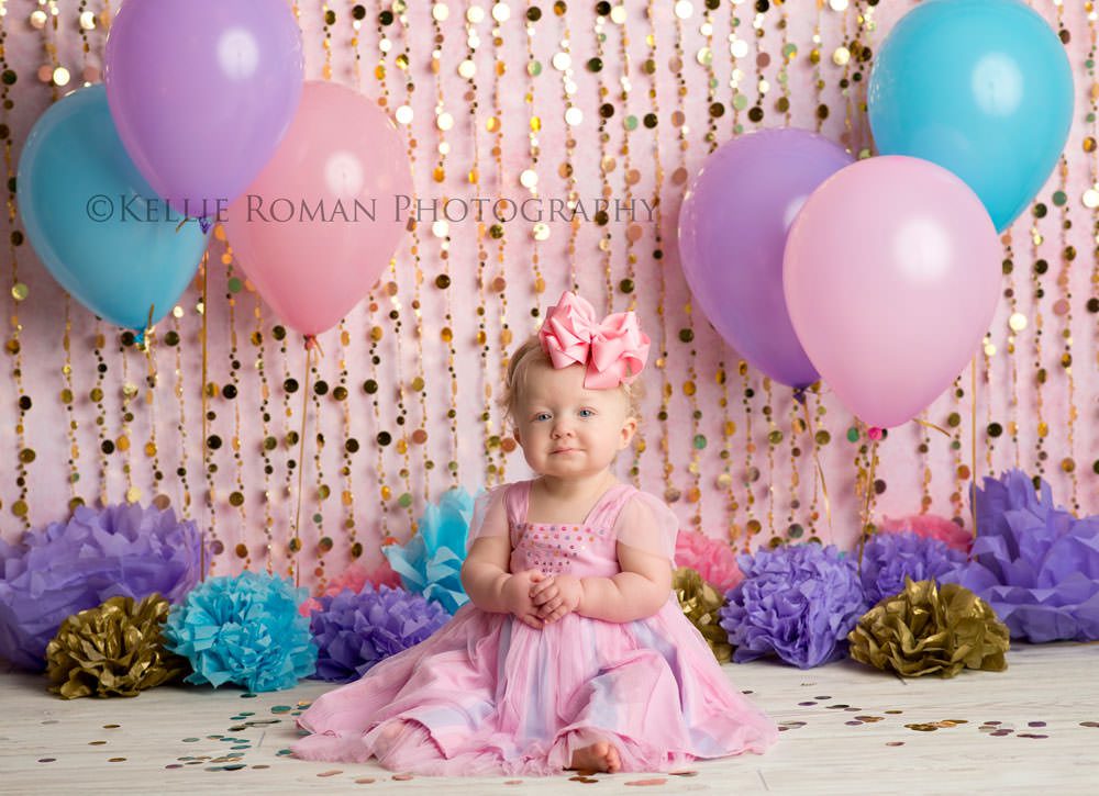 children photography one year old girl wearing a pink dress sitting infant of pink blue and purple balloons pom poms and gold beads