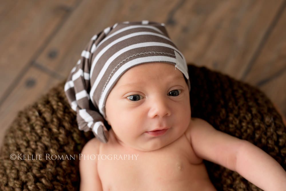 local milwaukee photographers newborn boy wide awake laying on top of brown blanket in bucket with striped hat on