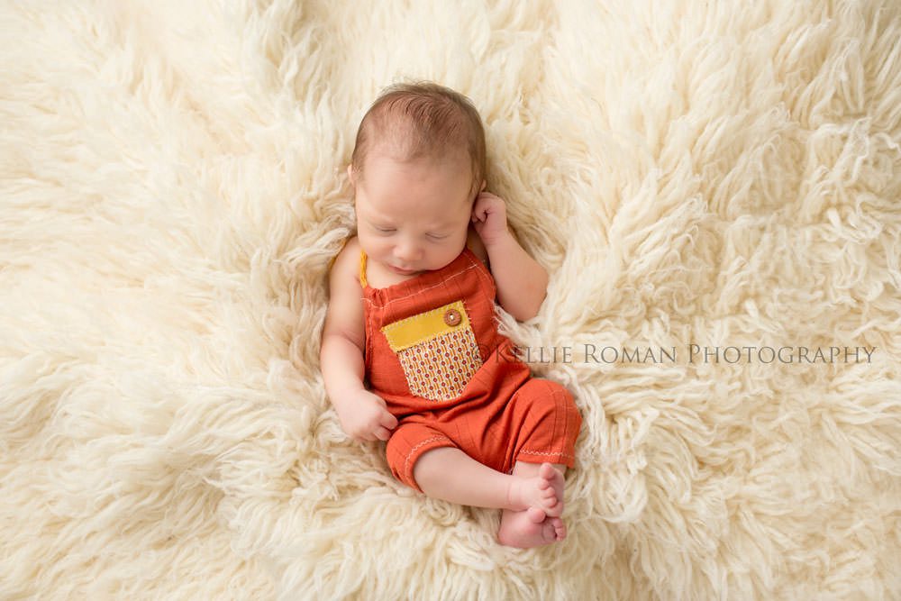 local milwaukee photographers newborn boy in orange romper with yellow pocket on front sleeping onto of ivory fur rug 