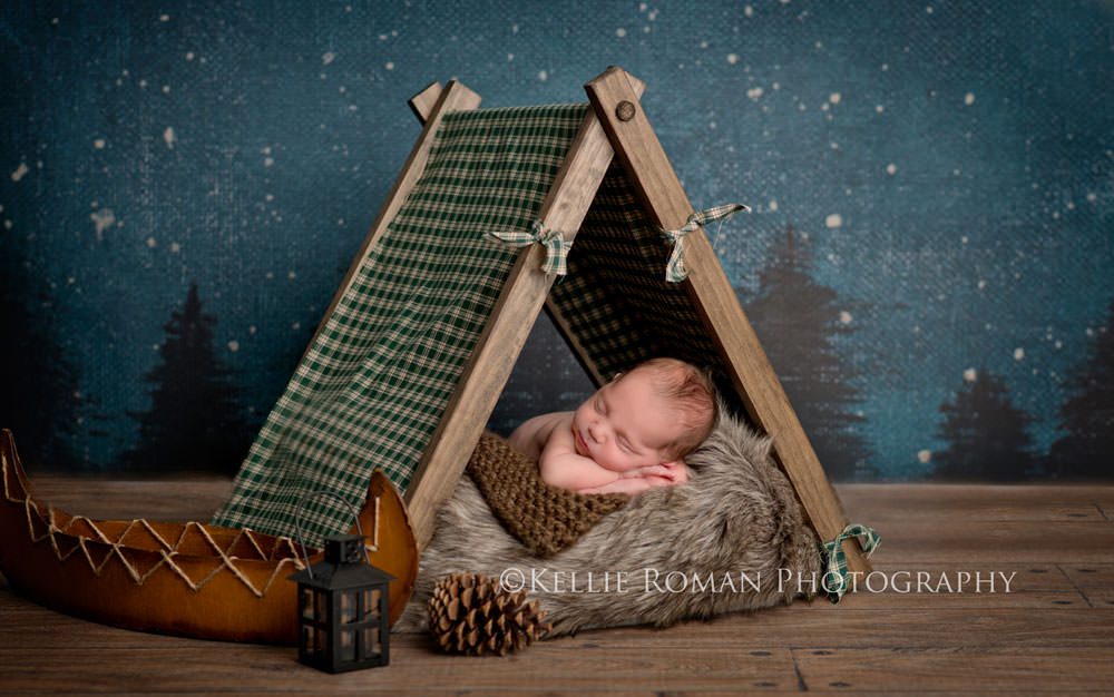 local milwaukee photographers newborn boy sleeping on belly under a green tent he has a canoe pinecone and lantern next to him there is a night sky backdrop with stars and pine trees behind him