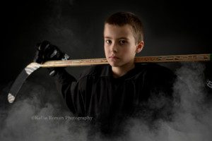 a boy is in a milwaukee photography studio. He has a black hockey jersey on, and black hockey gloves. He's holding his hockey stick with both hands behind his neck over his shoulders. He is looking into the camera with a serious face and there is white smoke all around him