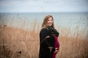 maternity. A women in a milwaukee county park near lake Michigan in Wisconsin is standing with her hands on her pregnant belly. She is standing in front of Lake Michigan in tall amber colored grass. she is wearing a maroon dress with a black sweater.