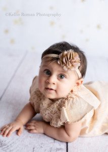 children studio a nine month old baby girl is in a milwaukee photo studio. She has a gold dress on with a headband and is laying on her belly. She's on a white wood floor with a white backdrop and gold stars behind her.