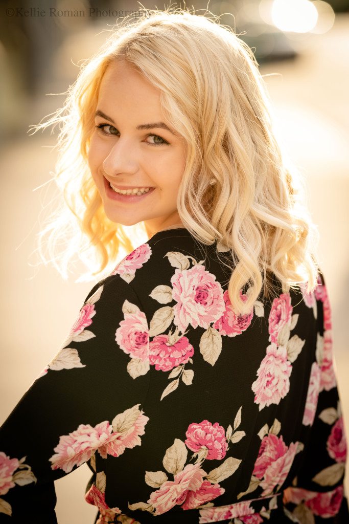 third ward senior session. a waukesha high school senior girl is in Milwaukee's third ward. the shot is a close up of her. she is faced the opposite way of the camera, has her hand on her hip and is turning while smiling at the camera. she's wearing a black romper with pink and white flowers on it. the sun is making her blonde hair glow.