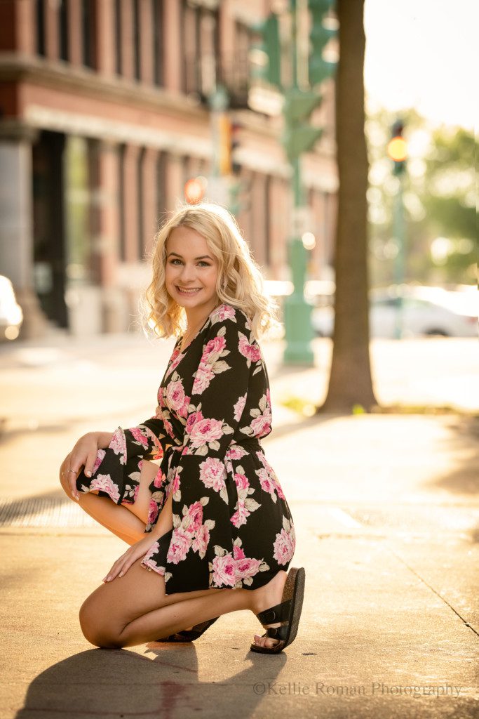 third ward senior session. a waukesha Wisconsin girl is in Milwaukee's third ward downtown. she's kneeling on a sidewalk with the gorgeous glowing sunset behind her. she is wearing black sandals with a black romper with pink and white flowers. there are cars and a city scape off in the distance.