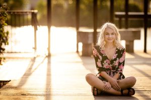 third ward senior session. a high school senior girl from waukesha is in downtown milwaukee sitting on a wood walkway. she is wearing sandals and a black romper with pink and white flowers on it. she is sitting criss cross, and the sun is glowing while setting behind her. there is a black metal structure behind her as well.