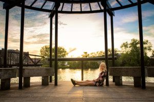 third ward senior session. A high school senior girl from waukesha is in Milwaukee's third ward for her senior session. she is sitting on a wood walkway under a metal structure. the sun is setting behind her. she is wearing a black romper with a floral pattern