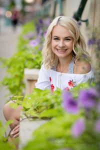 third ward senior pics. a waukesha senior girl is in the third ward in milwaukee having her senior pics taken. she's sitting on a step behind a pot of green and purple flowers. she's looking at the camera and smiling. she has on a white romper with red flowers, she has blonde hair and blue eyes.