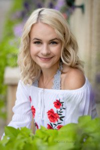 third ward senior session. a high school senior girl from waukesha is in downtown milwaukee for her senior pics. she's sitting on a step that you can't see. she's behind a bunch of green and purple flowers. she's wearing a white romper with red flowers. the girl has blonde hair and blue eyes.