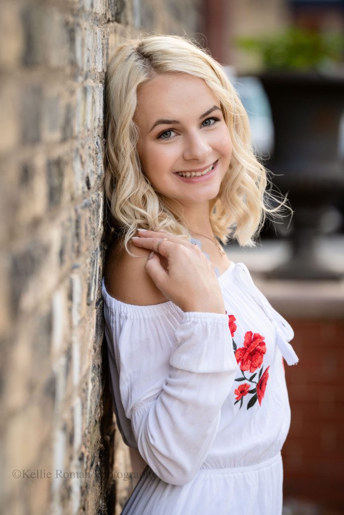 third ward senior session. a high school senior girl is in download milwaukee. She has her back against an old brick building and is looking at the camera. she has her hand up on her shoulder, and is wearing a white dress. she has blonde hair and blue eyes.