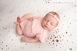 newborn girl in milwaukee photography studio. newborn baby is wearing a light pink romper with a light pink bow headband. she's sleeping on her back onto of a white blanket with gold stars.