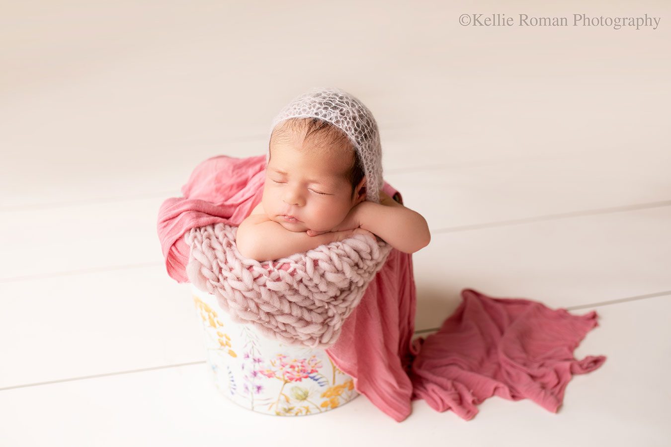Newborn baby girl is in Milwaukee Photographer studio. baby girl is sleeping upright in a floral bucket. newborn has her arms under her chin. she has a light pink bonnet on and the bucket is stuffed with light pink and dark mauve fabrics.