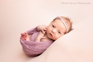 newborn Milwaukee Photographer. a baby girl is awake with her eyes open swaddled in a purple wrap laying on her back with her hands and feet sticking out. she has a light purple bow headband on and is laying on top of a light pink fabric.