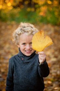 Young boy with blonde curly hair is outside in the fall. He is in the woods with trees of orange and yellow leaves around him. he has dark grey sweater on and blue eyes.