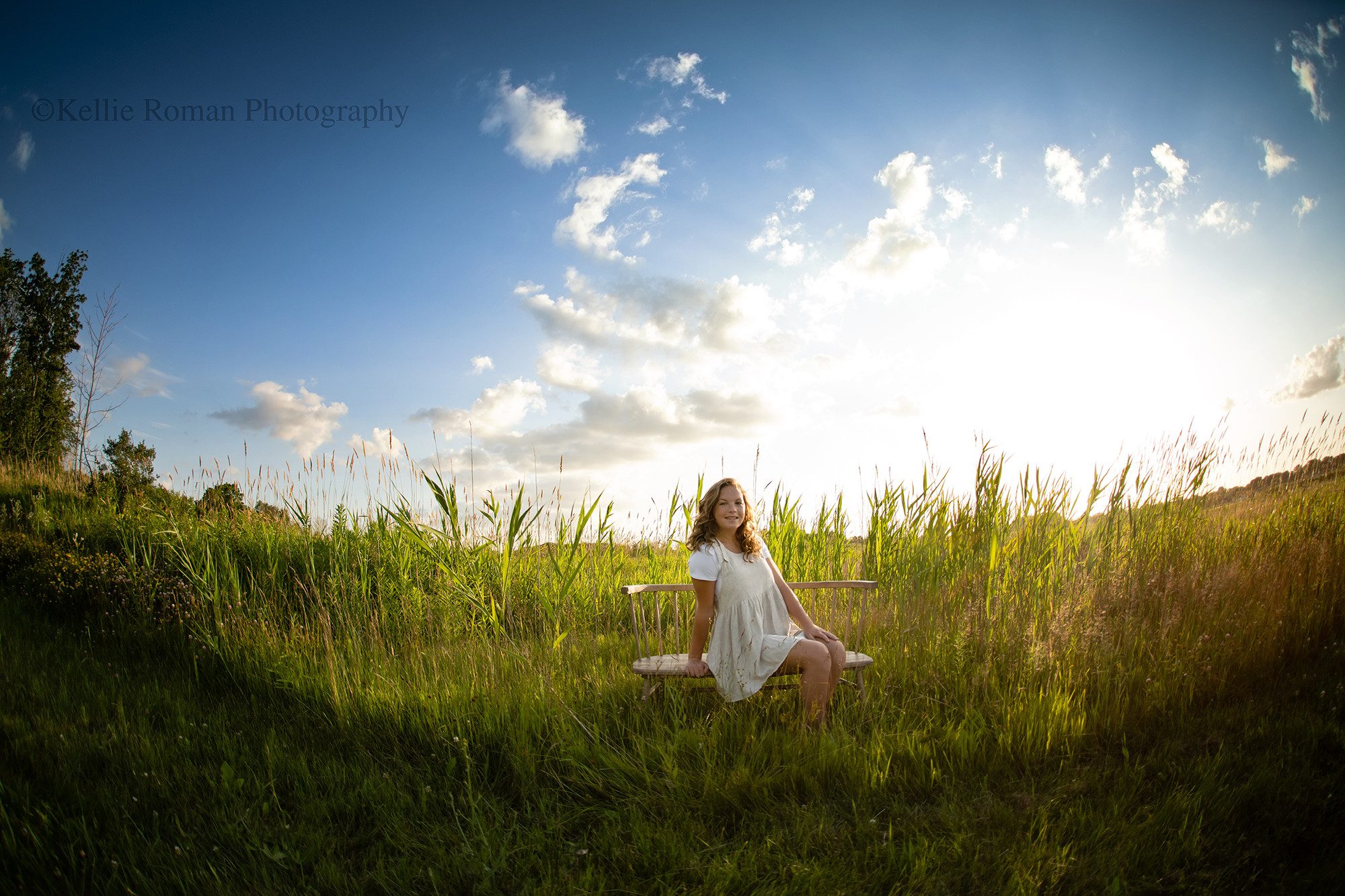 Milwaukee senior pics. A high school senior from Oak Creek is sitting on a wood bench in tall green grass in a park. The sunlight is shining from behind her, and the sky is blue with a few puffy clouds.