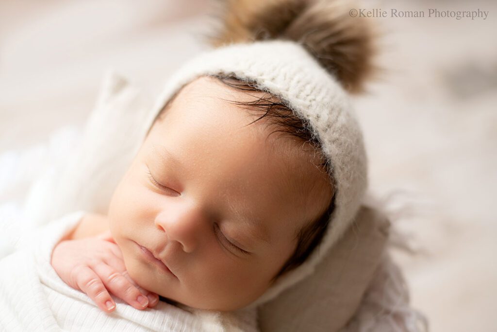 Newborn baby boy is sleeping while swaddled in a cream blanket with one hand sticking out of it. he has a cream colored bonnet on with a light brown poofy fur on top.