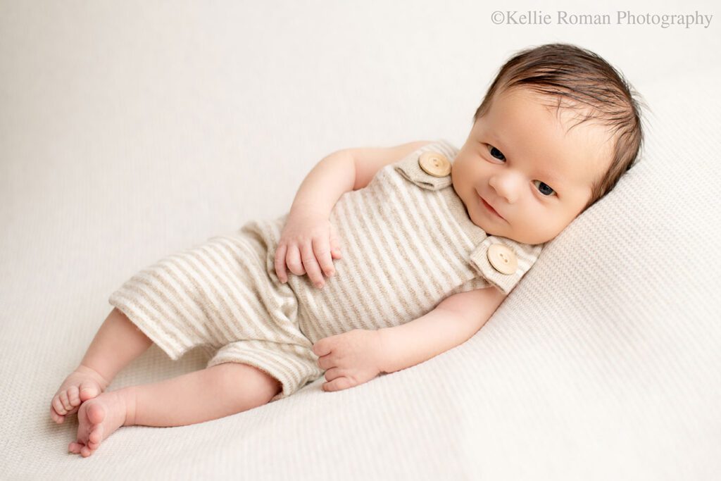 Newborn boy is wide awake in milwaukee photo studio. he's laying on his side on a cream colored fabric, with a cream and beige striped romper with big wood buttons. he has brown hair and is smiling.