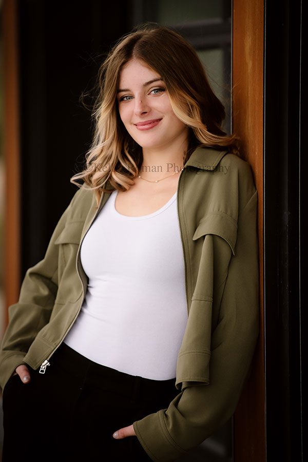 milwaukee senior photographers. high school senior girl wearing an olive green jacket and white tank top is leaning against a wall in oak creek park. 