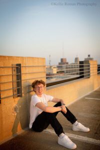 high school senior boy on the top level of a Downtown Milwaukee parking garage. he's sitting with back against concrete with buildings in the background with the sky. he has dark jeans on with white shoes and a white shirt. he's smiling.