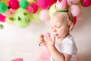 milwaukee cake smash pictures. image of a one year old girl looking at her hand that is covered in frosting. she has a pink glitter crown on with a watermelon. the backdrop is bright pink and green watermelon balloons.
