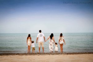 milwaukee family photographers. a family of 6 is at the beach in milwaukee. they are holding hands while looking out onto the waters of Lake Michigan. the family is wearing colors of creams, ivory, and florals. the water and sky are both very blue.