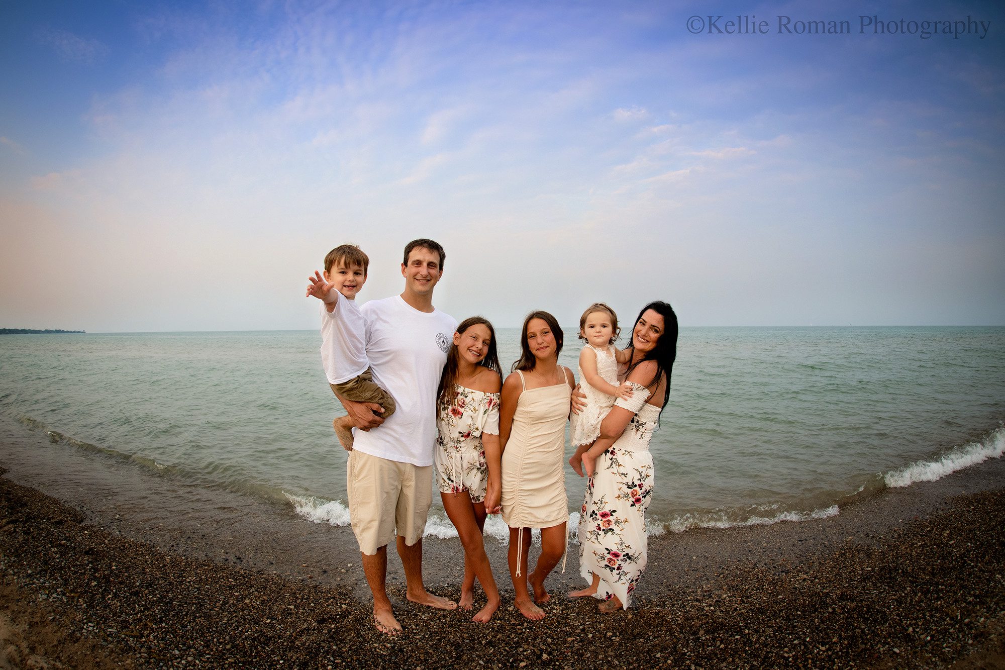 milwaukee family photographer. a family of 6 with 4 children are standing on a beach along Lake Michigan. the family is wearing shades of cream and floral. the sky is bright blue, and the water is teal blue.