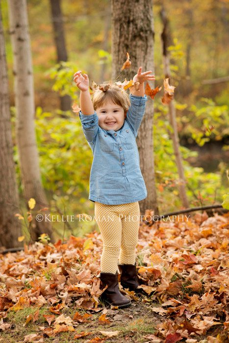 milwaukee child photographer little girl throwing leaves up in the air smiling she's standing in a park with trees surrounding her wearing brown boots yellow leggings and a denim shirt