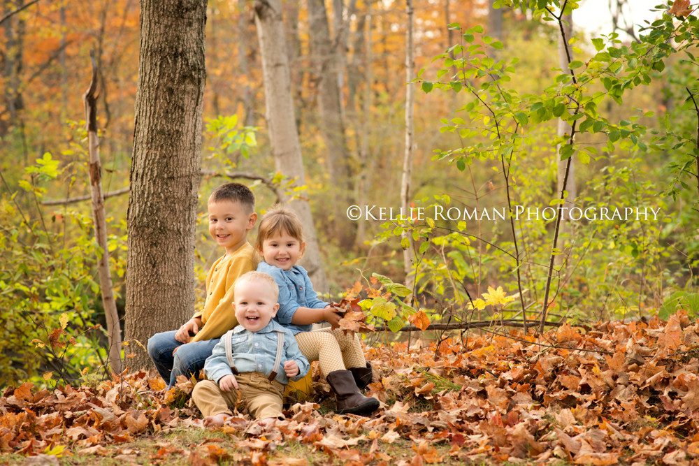 milwaukee child photographer three kids sitting on a yellow bucket in a bed of fall leaves they are wearing shades of yellow denim and khaki