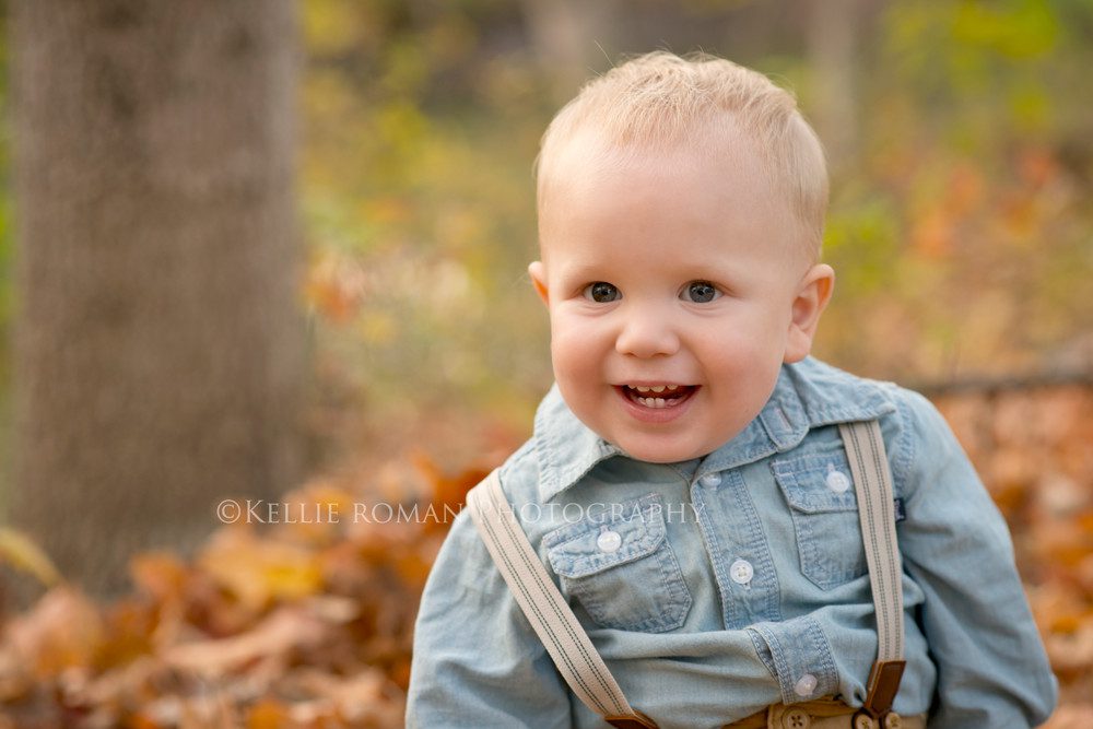 milwaukee child photographer boy sitting in the woods on a bed of yellow and orange fall leaves smiling very big wearing a denim shirt and suspenders
