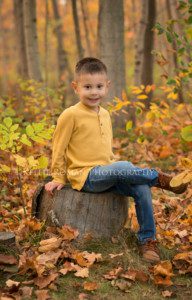 milwaukee child photographer boy sitting in the woods on a stump with yellow, and orange fall leaves around him he's wearing a yellow shirt jeans and brown boots