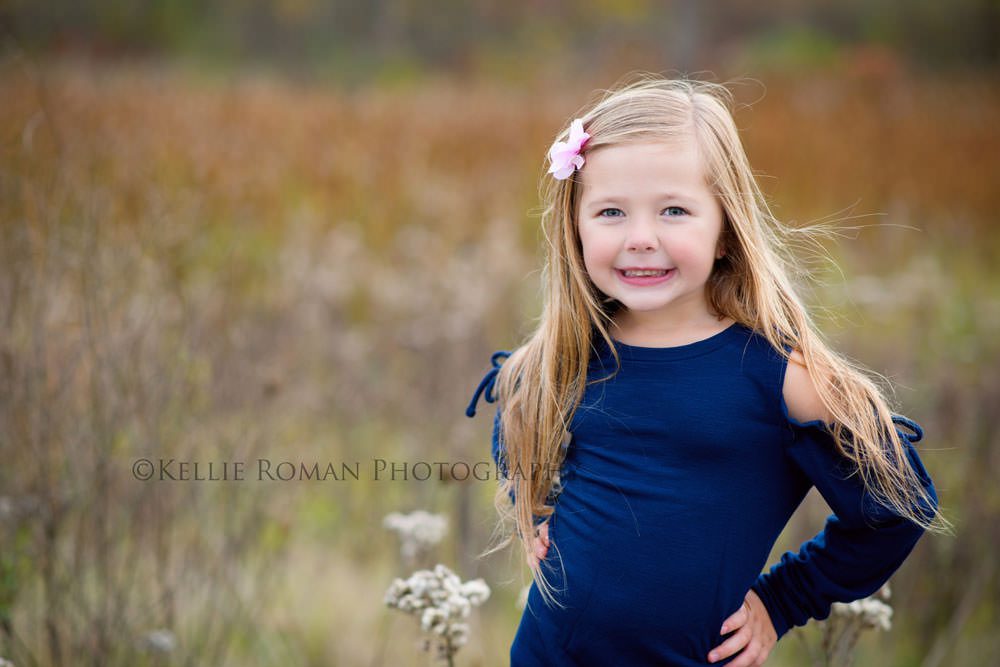 outdoor family photographer little girl standing outside in park with her hands on her hips she has long hair and is wearing a navy dress while smiling at the camera