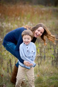 outdoor family photographer mother playing with son with her arms around him they are both laughing and smiling very big outside in a park mother is wearing navy shirt with jeans and son has a grey sweater on