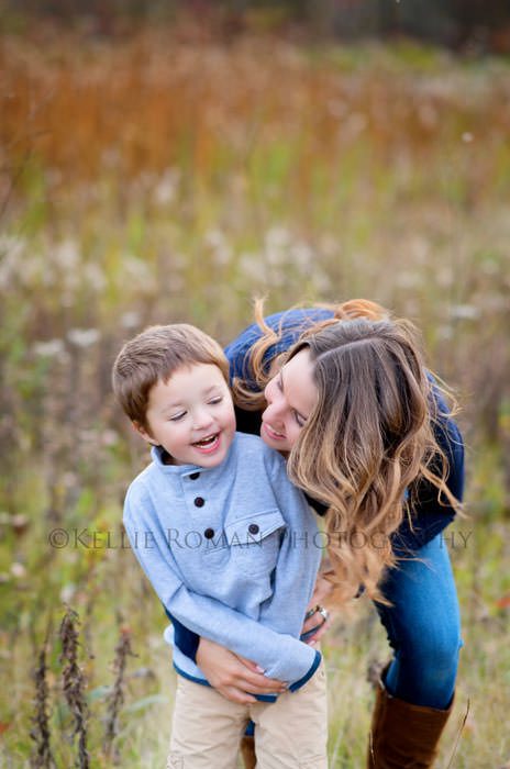 outdoor family photographer mother hugging son from behind they are both smiling very big while looking at each other they are outside in a park during the fall