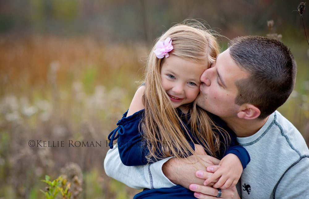 outdoor family photographer father hugging daughter from the side while giving her a kiss on the cheek they are outdoors in a park daughter is wearing navy and father has a grey shirt on