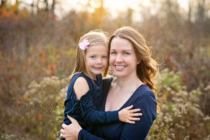 outdoor family photographer mother holding daughter in park with fall colored leaves around them they are both wearing navy and the sunshine is coming through their hair