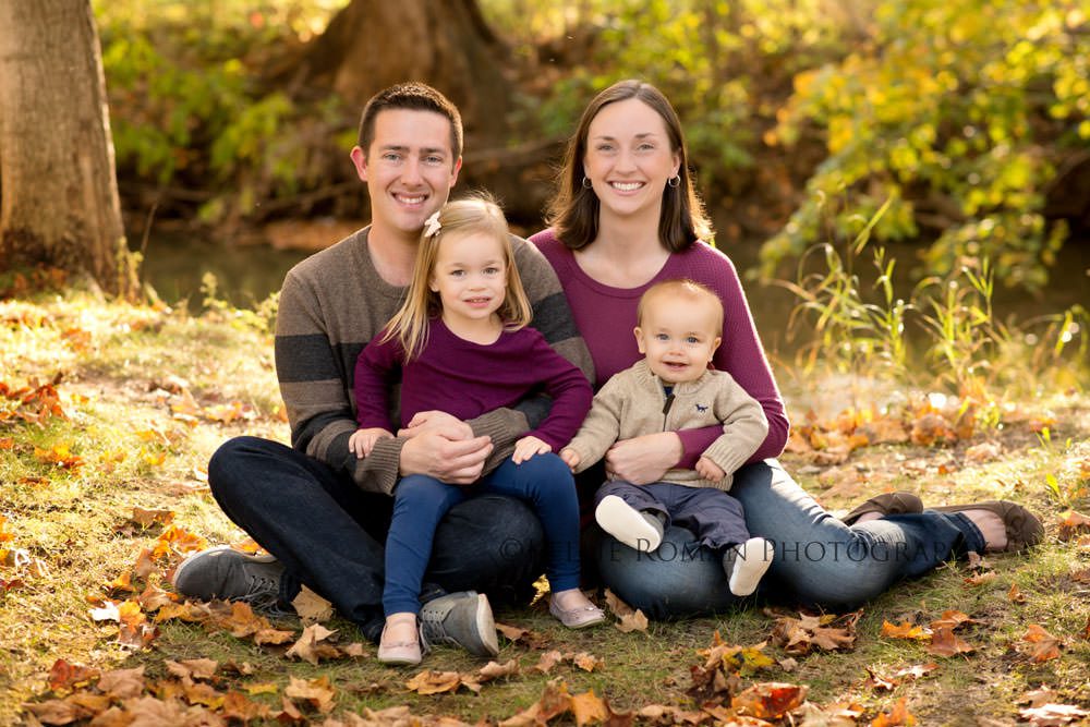 fall sunlight family photographer family with two kids a boy and a girl sitting on the ground in a park surrounded by orange and yellow leaves they are smiling and looking at the camera
