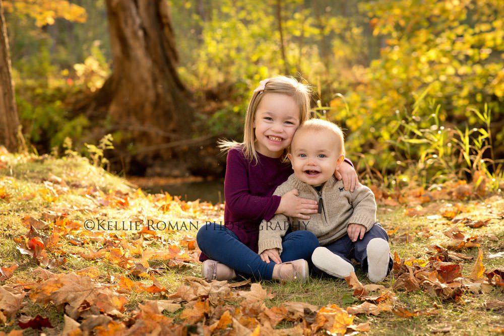 fall sunlight family photographer two kids a brother and sister sitting on the ground in a park surrounded by yellow and orange leaves they are smiling at the camera