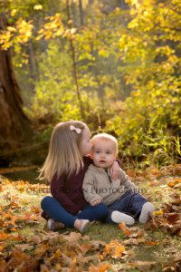 fall sunlight family photographer two kids one boy and one girl sitting on the ground in park with fall leaves around them the little girl is kissing her brother on the head