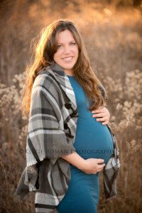 maternity pics in november a women who's pregnant standing in a field of tall brush holding her pregnant belly she's wearing a tight teal dress and a black and white sweater she is looking into the camera and the sun is shining through her hair