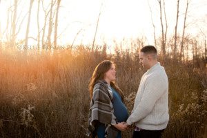 maternity pics in november a husband and wife who is expecting standing outside in a field of tall grass the sun is creating sun flair and they are looking at each other while holding hands