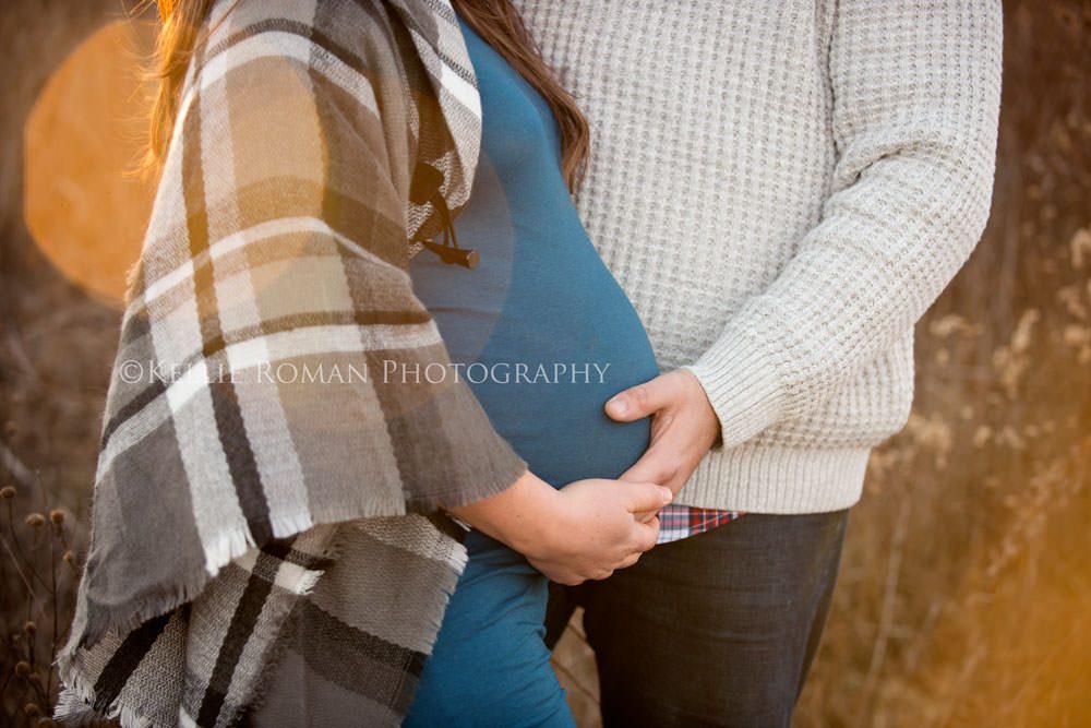 maternity pics in november a close up shot of the belly of a pregnant woman and her husband with their hands on her belly they are outside in a park with sun flair in the image