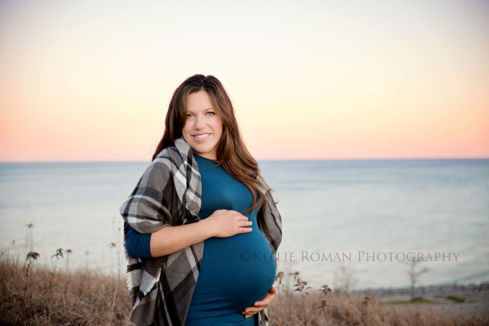 maternity pics in november a pregnant woman waring a teal dress and plaid sweater holding onto her belly she's in front of lake michigan and is looking at the camera